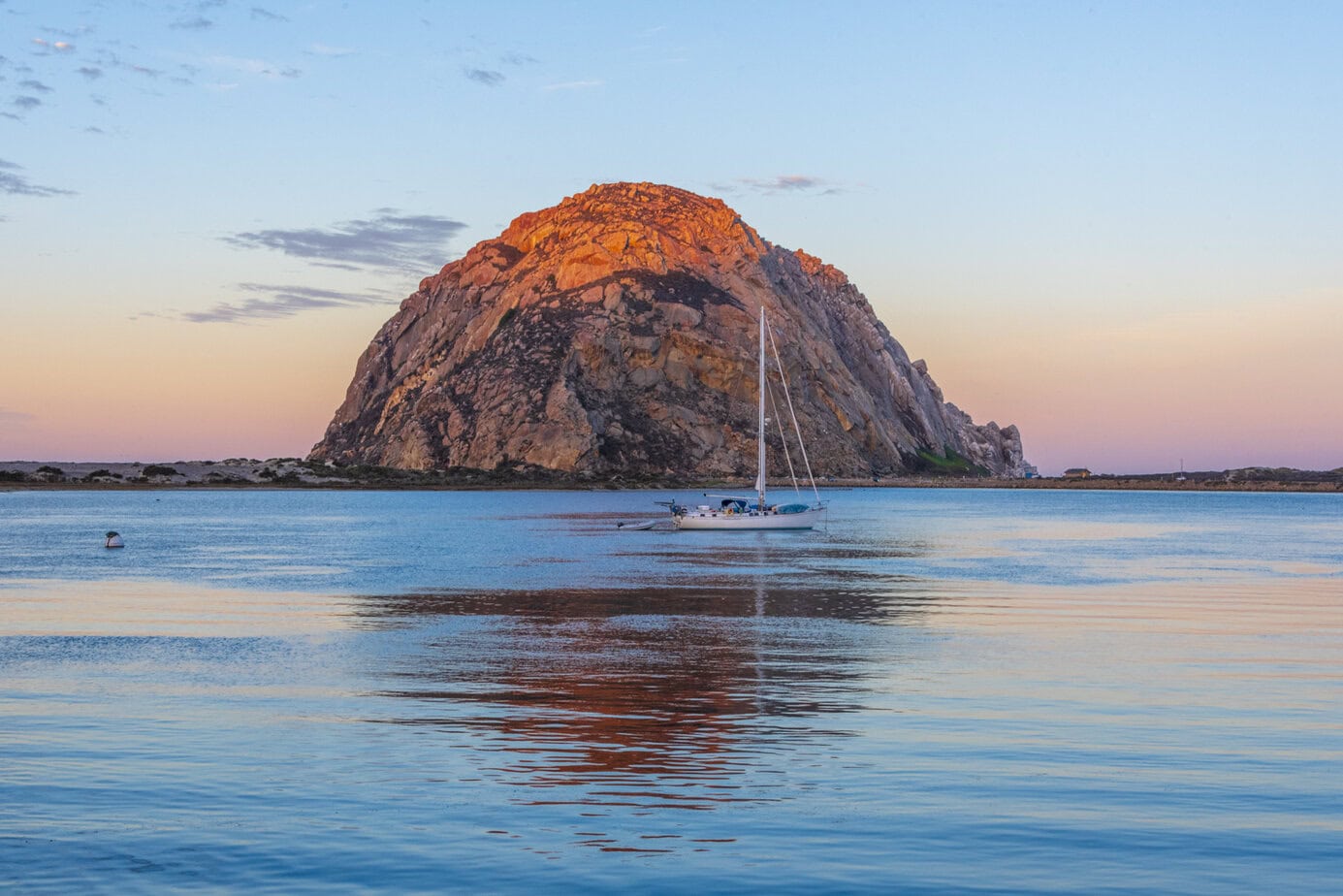 Morro Rock bathed in warm sunset light, with a sailboat drifting on calm waters in Morro Bay, California.