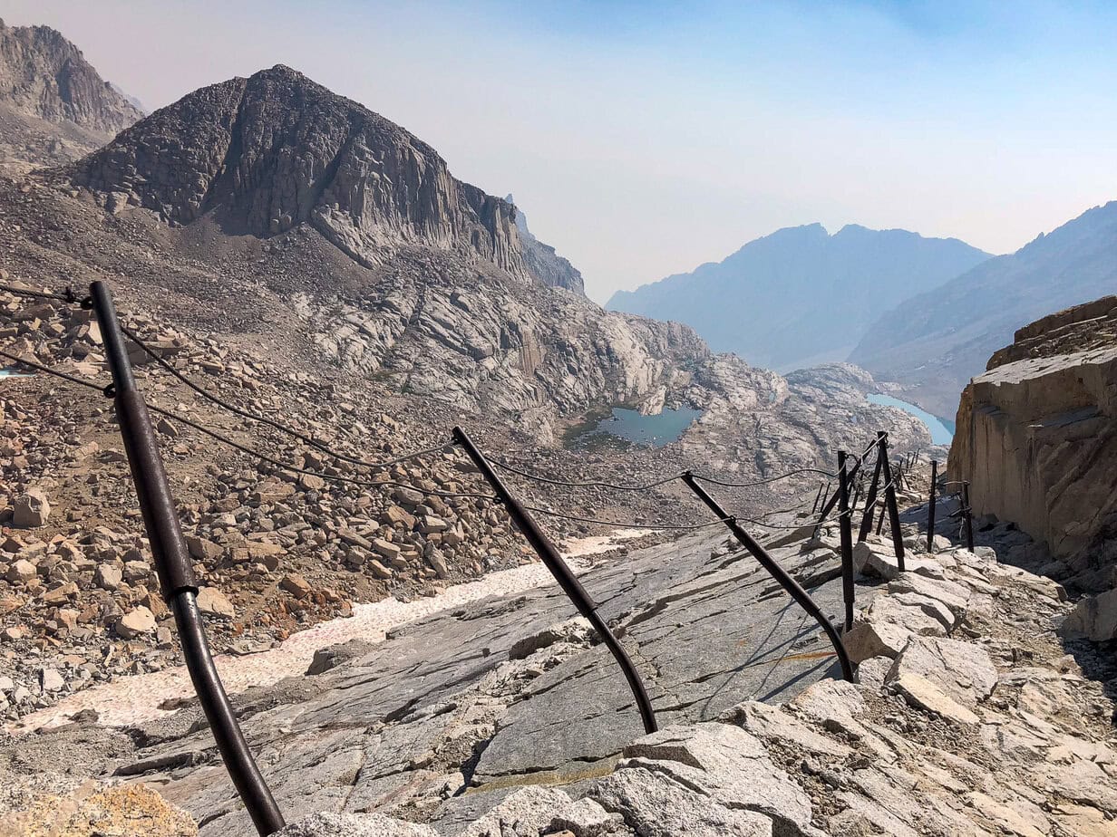 The exposed cable section along the Mount Whitney Trail, featuring safety cables anchored to granite rock above high alpine terrain.