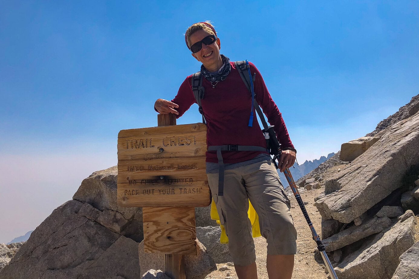 Hiker standing by the wooden Trail Crest sign on the Mount Whitney Trail, marking the highest point on the John Muir Trail and the entrance to Sequoia National Park.