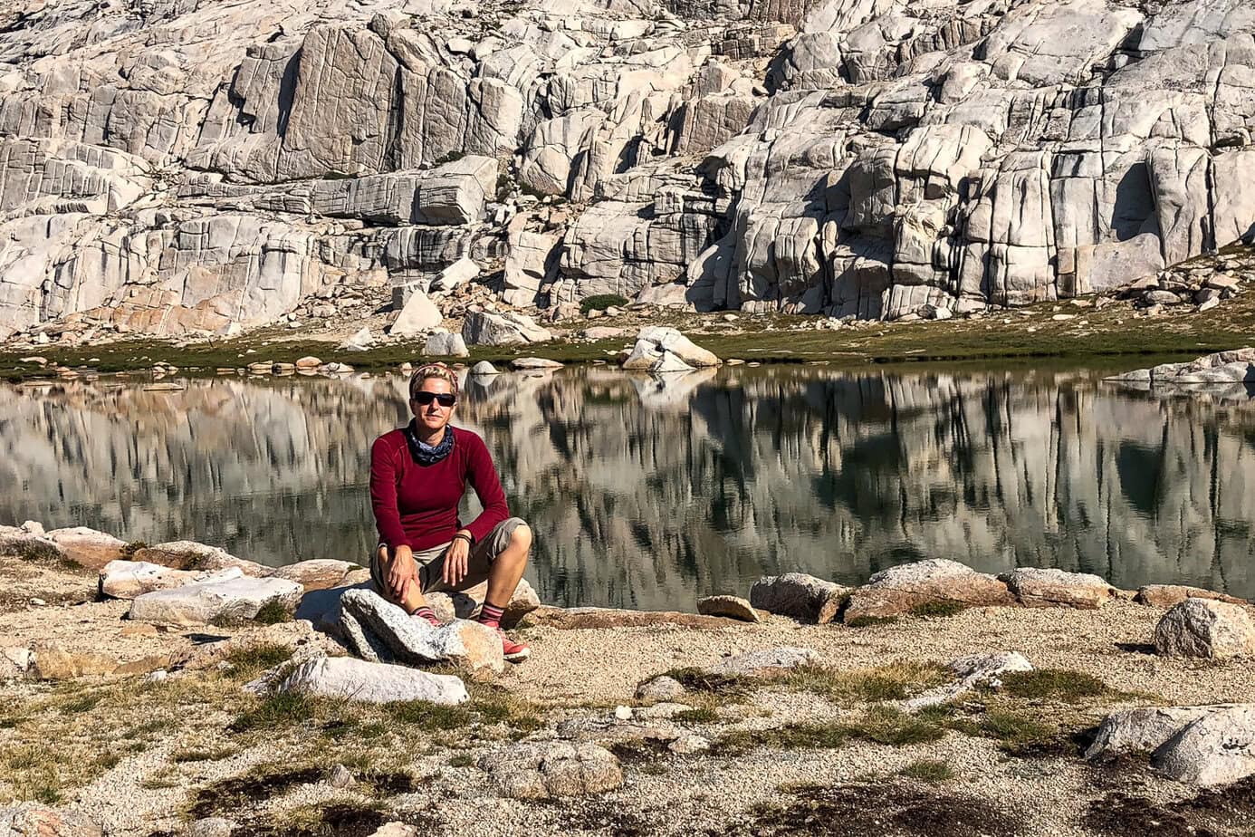 Hiker resting beside a high-altitude alpine lake at Trail Camp on the trail, with jagged granite cliffs reflected in the water.