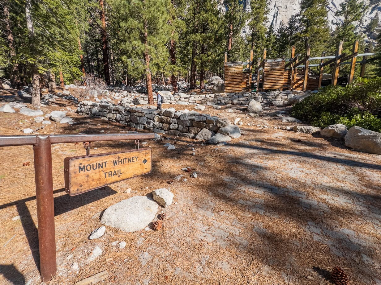 Mount Whitney Trailhead sign at Whitney Portal, framed by forest, stone walls, and information stations—marking the start of the iconic Sierra Nevada hike.