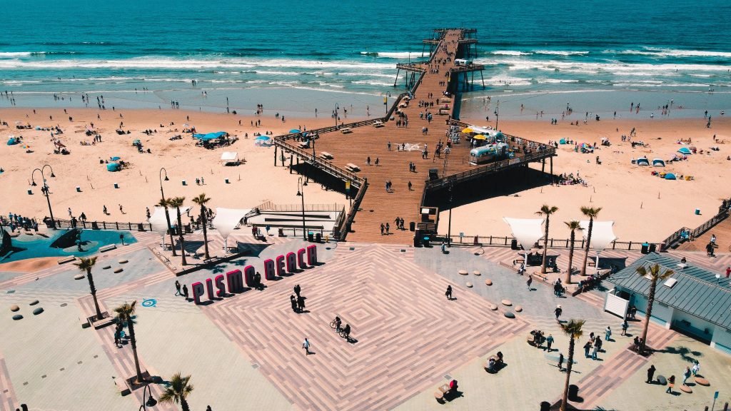 Pismo Beach Pier seen from above, showing the boardwalk, Pismo Beach sign, and Pacific Ocean waves on California’s Central Coast.