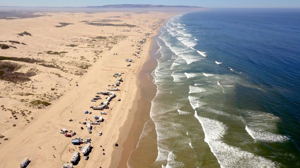 Aerial view of the Pismo Beach dunes with vehicles and campers lined up along the shoreline beside breaking Pacific waves.