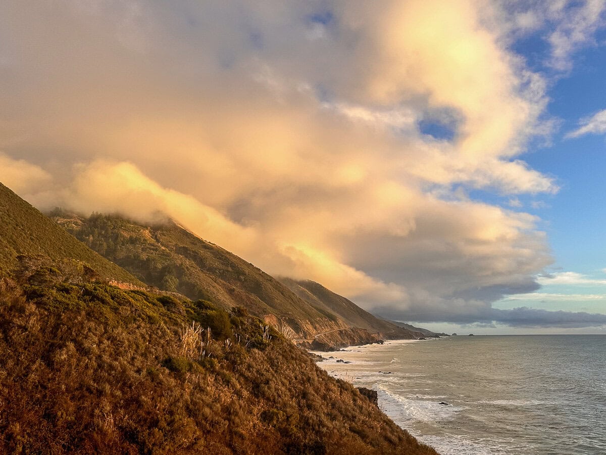 Sunlit clouds drifting over rugged Big Sur cliffs and the Pacific Ocean along Highway 1 at sunset