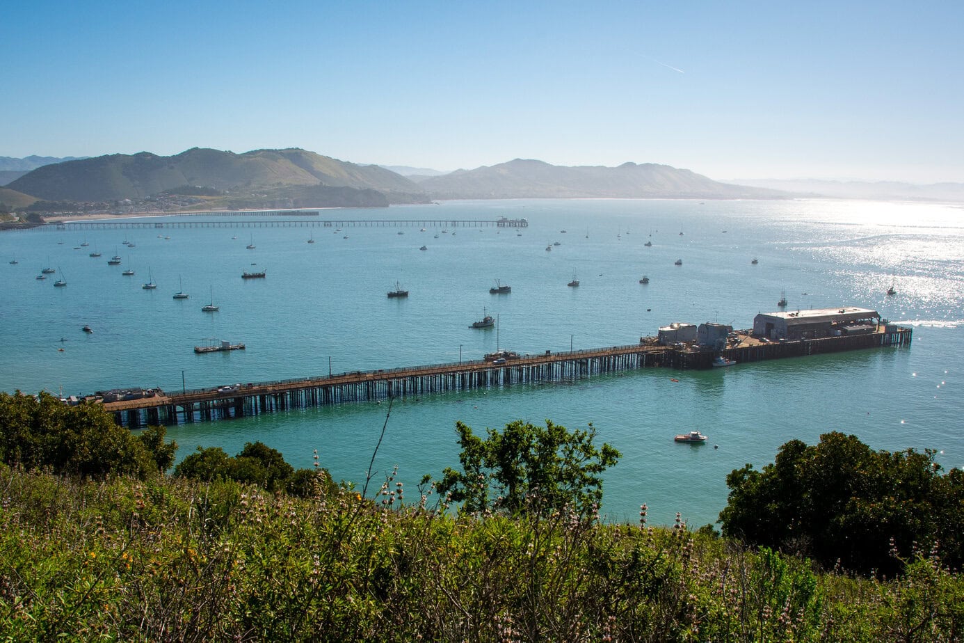 Elevated view of Avila Beach’s fishing pier surrounded by anchored boats, with the Cal Poly Pier in the background and rolling hills framing the Pacific horizon.