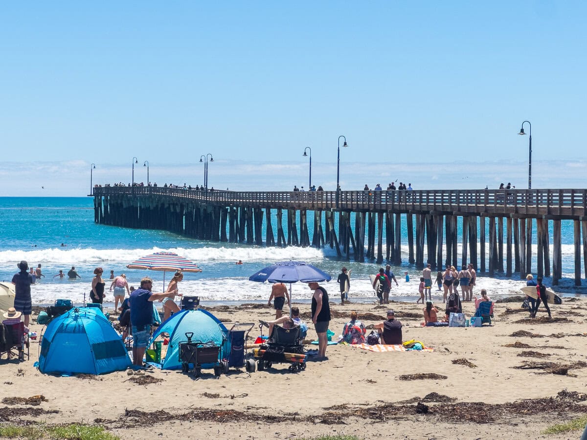 Sunny view of Cayucos State Beach and the pier with umbrellas and beachgoers.