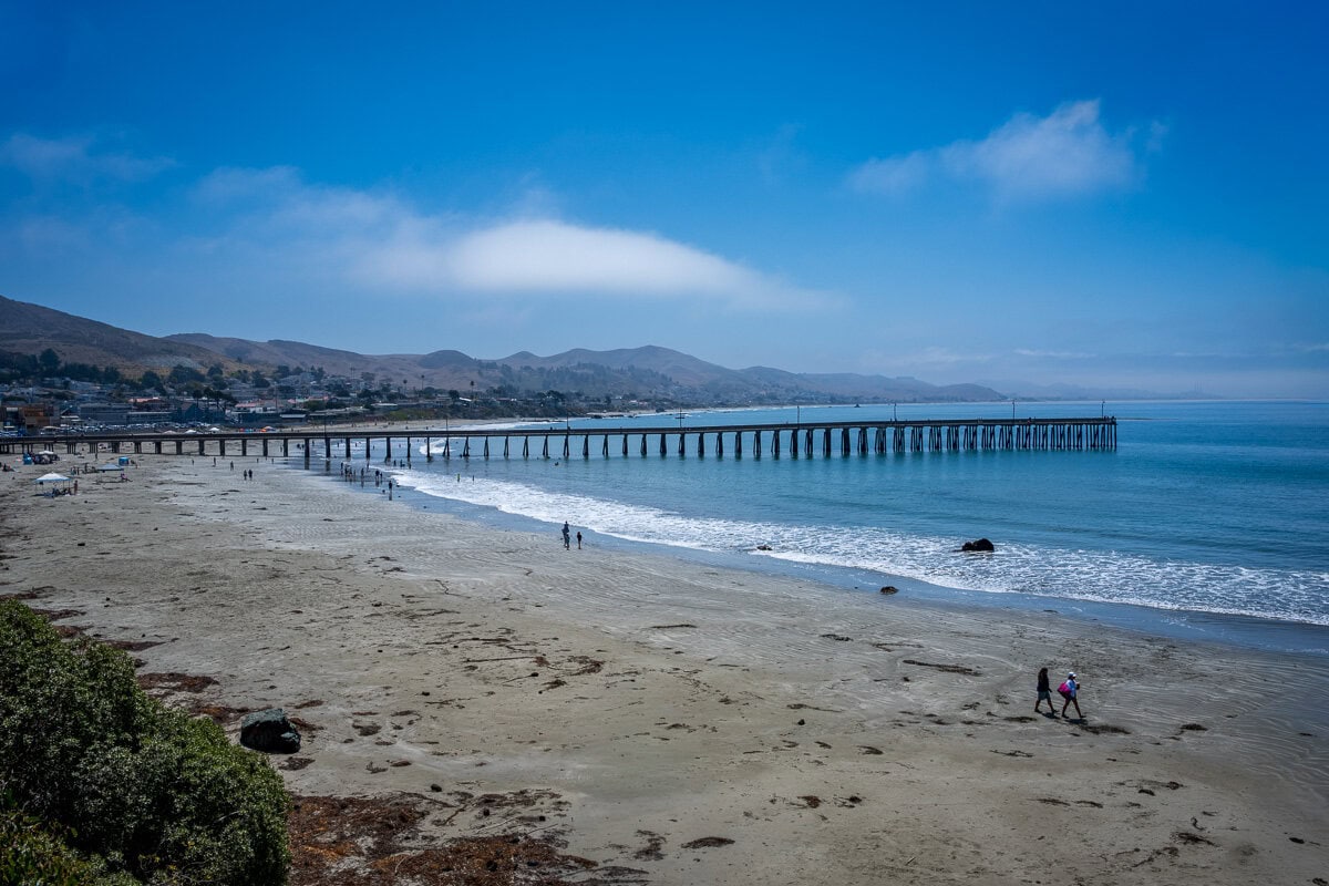 Wide view of Cayucos Pier stretching across the beach beneath blue sky and coastal fog