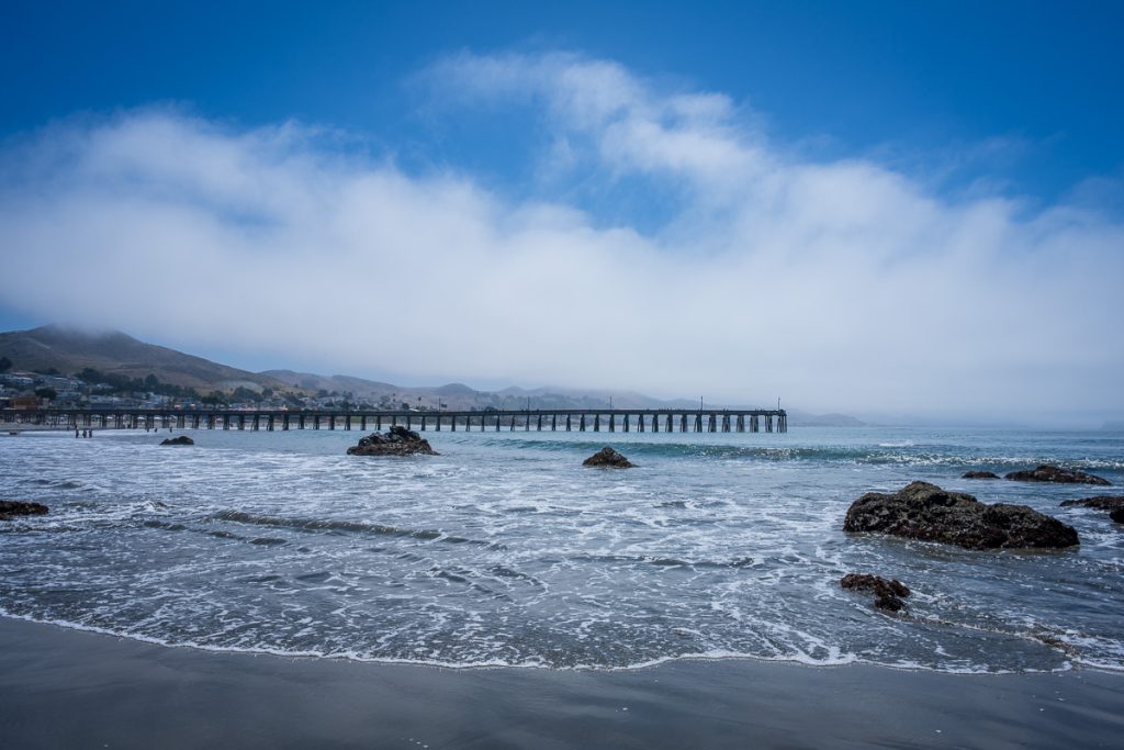 Rocky shoreline and Cayucos Pier beneath morning fog and a patch of blue sky