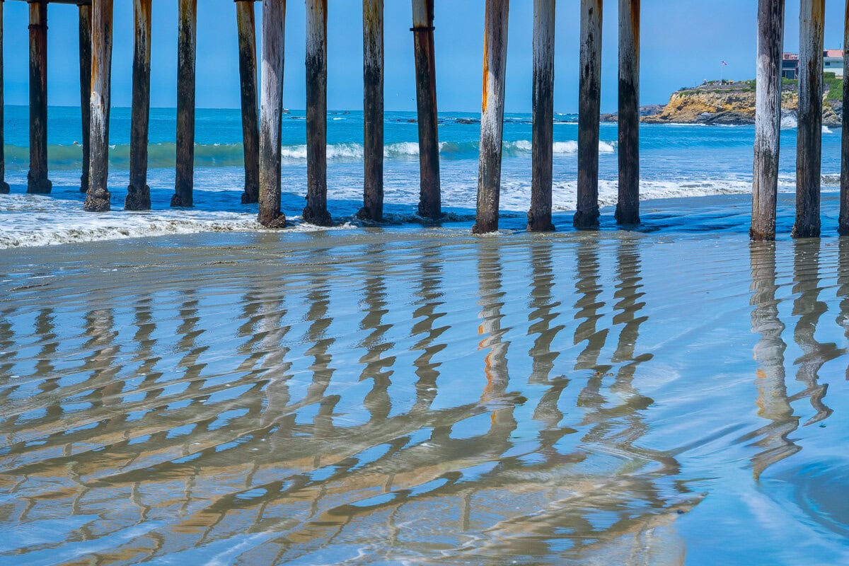 Wooden pilings of Cayucos Pier reflected in shallow water on the sand