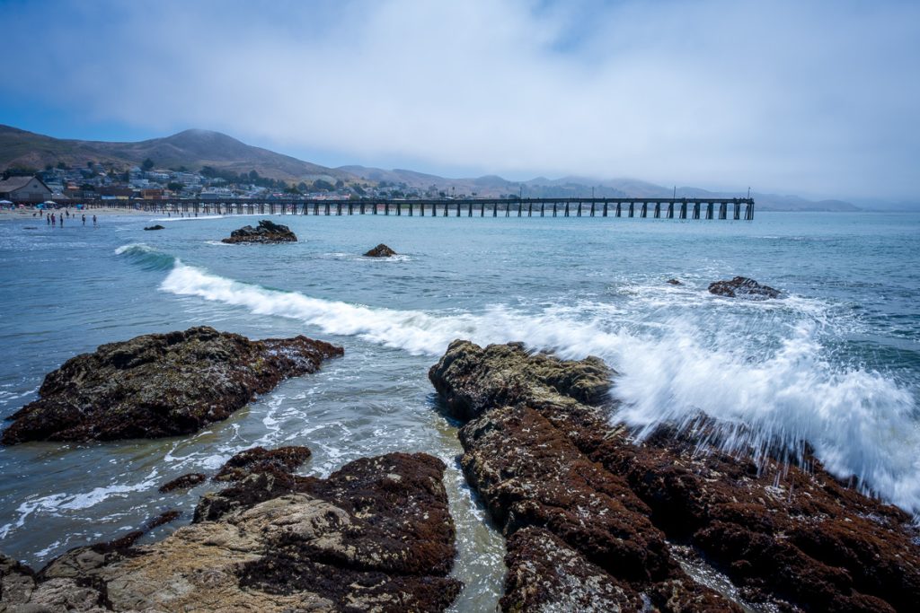 Waves breaking over shoreline rocks with the long pier visible in the distance