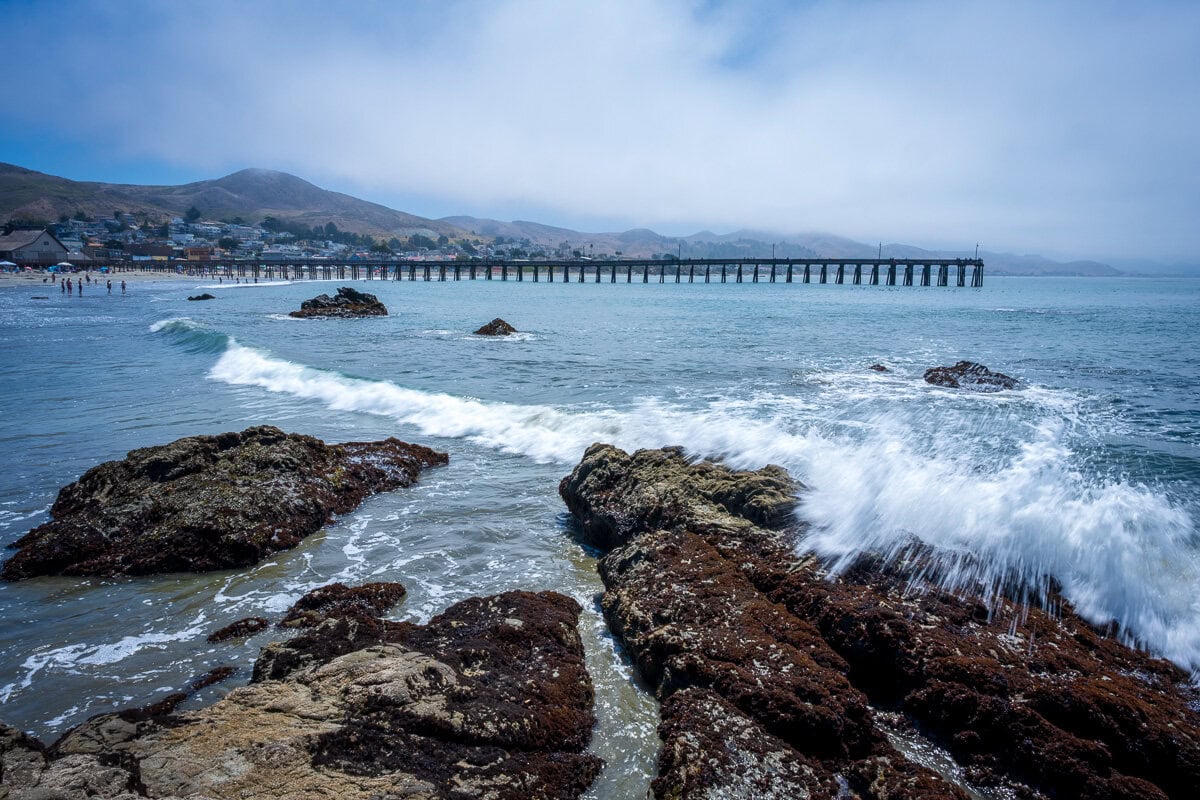 Waves breaking over shoreline rocks with the long pier visible in the distance