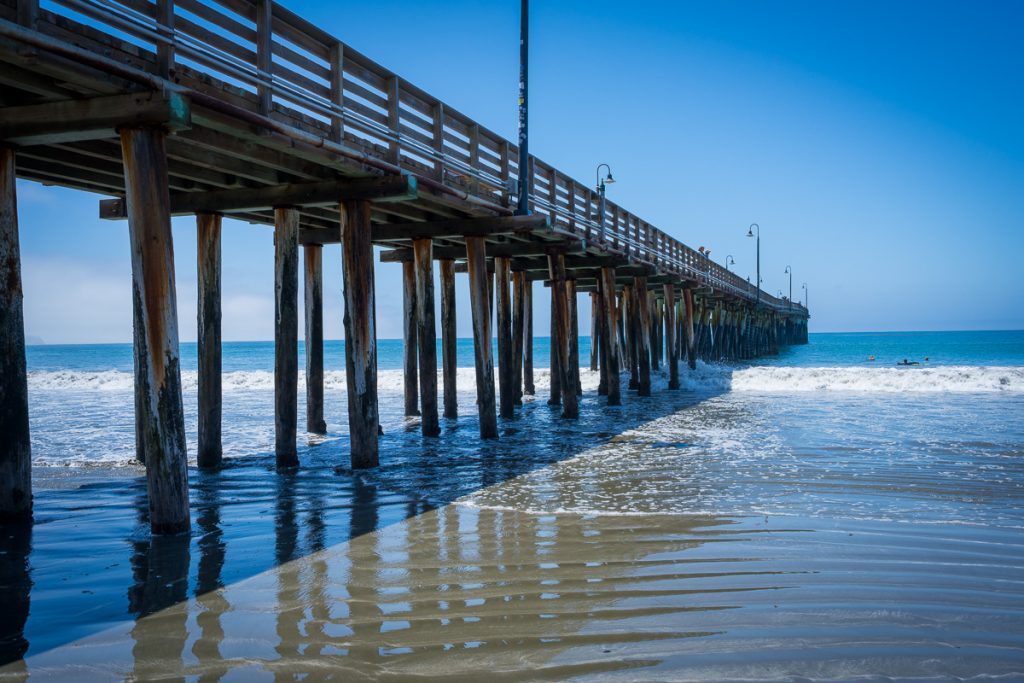 Angled view beneath Cayucos Pier showing wooden pilings reflected on wet sand